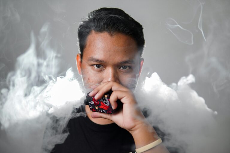 Young man exhaling large vape cloud while holding a decorative vape device, wearing a black shirt