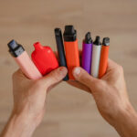 Two hands holding a collection of colorful disposable vapes in pink, red, black, orange, purple, and white.