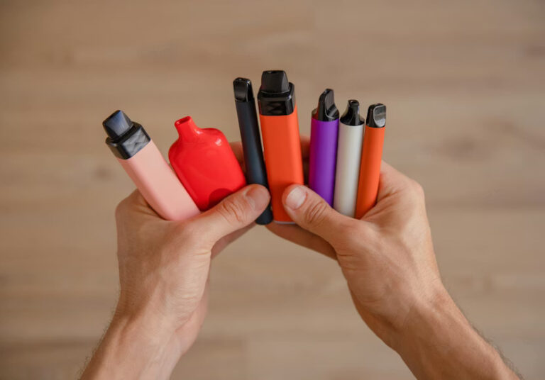 Two hands holding a collection of colorful disposable vapes in pink, red, black, orange, purple, and white.